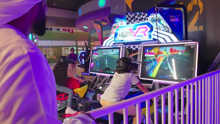 طفلان عربيان خليجيان اماراتيان يرتديان الزي الكاجوال يلعبان لعبة سباق الدراجات النارية التفاعلية داخل مركز ألعاب إلكترونية، أجواء حماسية وتجربة تنافسية ممتعة، ألعاب سباق إلكترونية للأطفال