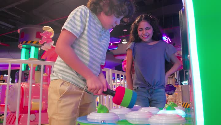 ايماءات السعادة والمرح داخل صالة ألعاب إلكترونية، مفهوم الترفيه والتسلية للأطفال، ولدعربي خليجي اماراتي يرتدي الزي الكاجوال يستخدم المطرقة في لعبة ضرب الدمى التفاعلية برفقة اخته