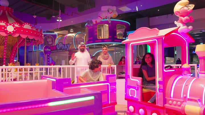 Expressions of happiness and fun inside an amusement center, two Emirati Gulf Arab children dressed in casual attire playing the amusement train game amidst a delightful family atmosphere, with parents participating in following the play experience.