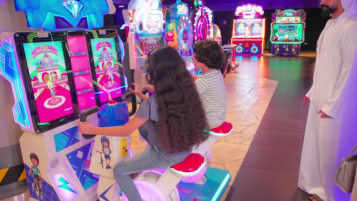 ايماءات السعادة و السرور، متابعة الاباء للأبناء وتعزيز روح المرح والتفاعل الأسري، قضاء أوقات ممتعة مع العائلة، طفلان عربيان خليجيان اماراتيان يرتديان الزي الكاجوال يلعبان لعبة سباق إلكترونية تفاعلية داخل صالة ألعاب