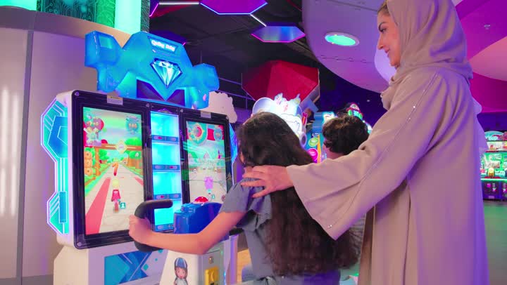 قضاء أوقات ممتعة مع العائلة، طفلان عربيان خليجيان اماراتيان يرتديان الزي الكاجوال يلعبان لعبة سباق إلكترونية تفاعلية داخل صالة ألعاب، متابعة الاباء للأبناء وتعزيز روح المرح والتفاعل الأسري، ايماءات السعادة و السرور