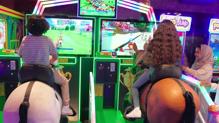 قضاء وقت ترفيهي ممتع داخل صالة ألعاب إلكترونية، تصوير من الخلف لطفلان عربيان خليجيان اماراتيان يرتديان الزي الكاجوال يستمتعان بلعبة سباق وركوب الحصان التفاعلية، متابعة الوالدين لابنائهم بايماءات السعادة و السرور