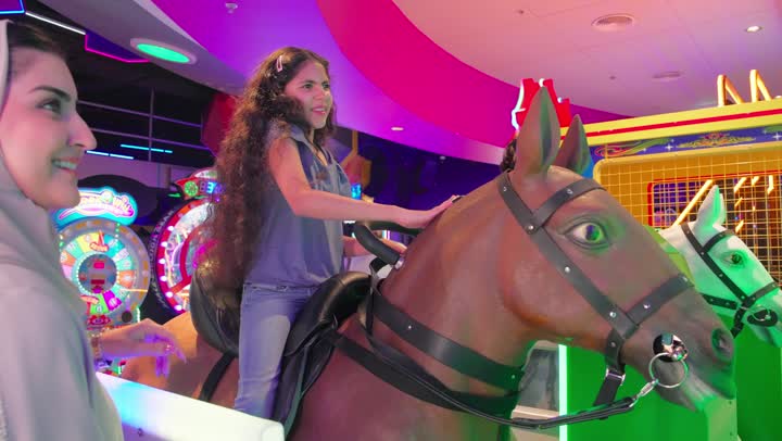 مفهوم الترفيه العائلي، بنت عربية خليجيه اماراتية ترتدي الزي الكاجوال تلعب لعبة ركوب الحصان الإلكترونية داخل مركز ألعاب حديث، مشاركة الأم لحظات السعادة والمرح