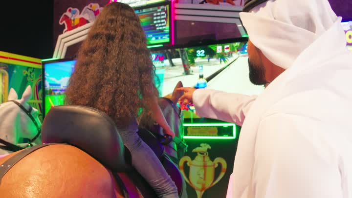 الاستمتاع بالألعاب الإلكترونية الحديثة، تصوير من الخلف لبنت عربية خليجية امارتية تجلس على لعبة ركوب تفاعلية وتشارك في اللعب بحماس، متابعة الأب لابنته بإيماءات التشجيع والدعم