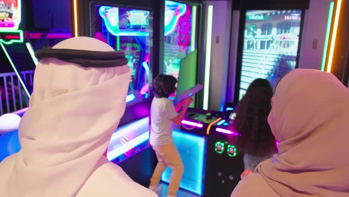 متابعة اللعب بإيماءات الإعجاب والاهتمام، أجواء ترفيهية داخل مركز ألعاب عصري، تصوير من الخلف لطفلين عربيين خليجيين اماراتيين يرتديان ملابس كاجوال يلعبان لعبة تصويب إلكترونية تفاعلية أمام شاشة أركيد مضيئة برفقة والديهم