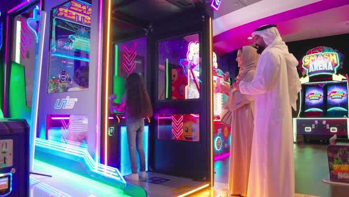 An entertaining atmosphere inside a modern game center, a shot from behind of two Emirati Gulf Arab children wearing casual clothes playing an interactive electronic shooting game in front of a brightly lit arcade screen, accompanied by their parents, following the game with gestures of admiration and interest.