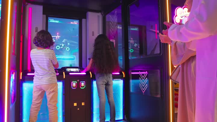 تنمية روح المشاركة والتفاعل بين الأطفال، قضاء وقت ممتع داخل صالة ألعاب إلكترونية، تصوير من الخلف لطفلين عربيين خليجيين اماراتيين  يرتديان ملابس كاجوال يلعبان لعبة تصويب إلكترونية تفاعلية أمام شاشة أركيد مضيئة برفقة والديهم ، تعابير السعادة و التشجيع