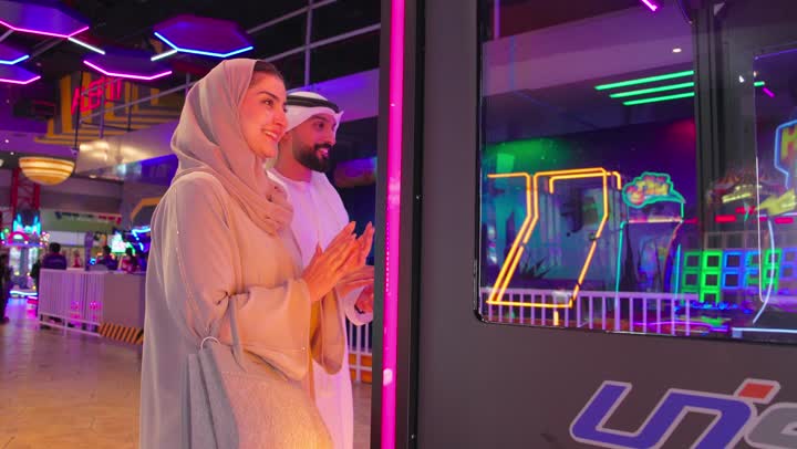 زوجان عربيان خليجيان اماراتيان يرتديان الزي التقليدي يقفان أمام إحدى الألعاب التفاعلية يقومان بالتصفيق ، تعابير السعادة و السرور، متابعة اللعب بإيماءات الإعجاب والاهتمام، أجواء ترفيهية داخل مركز ألعاب عصري