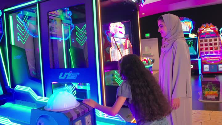 أم عربية خليجية اماراتية ترتدي العباءة و الحجاب ترافق ابنتها أثناء اللعب في لعبة البولينج الإلكترونية، مشاركة الأطفال في الألعاب التفاعلية، قضاء وقت ممتع داخل صالة الألعاب، الاستمتاع بالألعاب الإلكترونية الحديثة
