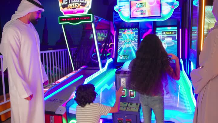 قضاء وقت ممتع داخل صالة الألعاب، رجل عربي خليجي اماراتي يرتدي الكندورة و الغتره  يشارك ابنائة اللعب في لعبة البولينج الإلكترونية، متابعة الاب للأطفال ودعمهم خلال التجربة، تعزيز الروابط الأسرية