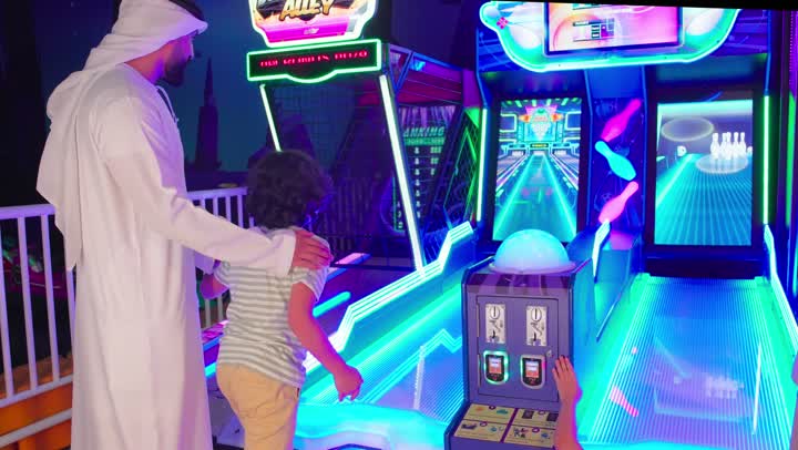 مشاركة عائلية في صالة ألعاب إلكترونية حديثة، تصوير من الخلف لأب عربي خليجي يرتدي اماراتي الكندورة و الغتره يرافق ابنة أثناء لعب لعبة البولينج الإلكترونية و يقوم بتشجيعة، قضاء وقت ممتع مع الأسرة، ايماءات  السعادة و السرور
