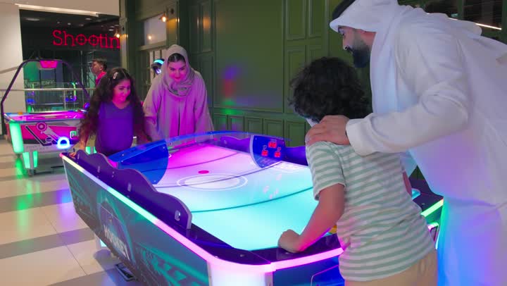 تعزيز الترابط الأسري من خلال الألعاب التفاعلية، قضاء لحظات ترفيهية مليئة بالحماس داخل مركز ألعاب إلكترونية، عائلة عربية خليجية اماراتية تشارك في لعب لعبة الهوكي الهوائي داخل صالة ألعاب ، تفاعل جماعي بين أفراد الأسرة أثناء اللعب بإيماءات المرح والتشجيع، قضاء وقت ممتع مع العائلة