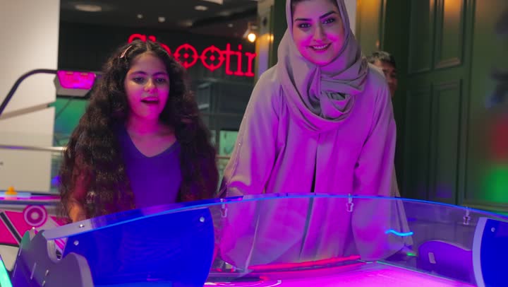 الاستمتاع بالألعاب التفاعلية، امرأة عربية خليجية اماراتية ترتدي العباءة و الحجاب برفقة طفلتها تشاركان اللعب على طاولة لعبة الهوكي الهوائي، لحظات حماس وتنافس ودي بين الأم وابنتها، قضاء وقت ممتع داخل مركز ترفيهي حديث، أنشطة عائلية تعزز التقارب الأسري