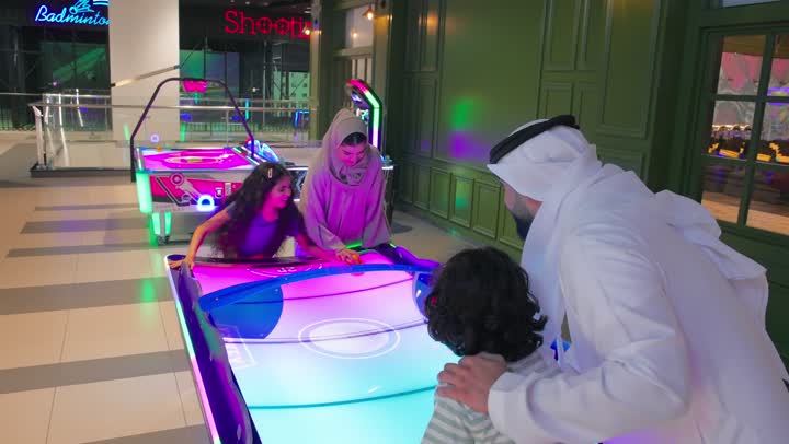 Spending exciting recreational moments inside an electronic games center, an Emirati Gulf Arab family participates in playing air hockey inside a game hall, with collective interaction among family members during the game through gestures of fun and encouragement, enjoying quality time with the family, and strengthening family bonds through interactive games.