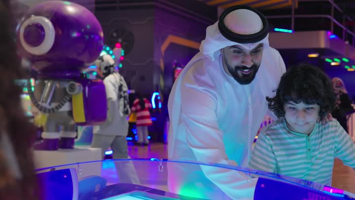قضاء لحظات الحماس داخل صالة ألعاب إلكترونية، أب عربي خليجي اماراتي  يرتدي الزي التقليدي يشارك ابنه الصغير اللعب على لعبة الهوكي، إيماءات التشجيع والدعم، تنمية روح التفاعل الأسري، الاستمتاع بالألعاب الإلكترونية الحديثة، تعزيز التواصل بين الآباء والأبناء داخل المراكز الترفيهية