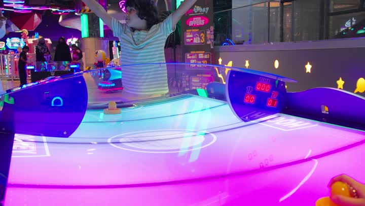 The interaction between family members, an Arab Gulf Saudi boy wearing casual attire raises his arms in gestures of joy after winning in air hockey inside a game hall, enjoying recreational and electronic games, having a good time, feelings of happiness and excitement, recreational activities for children.