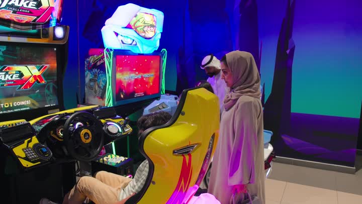 Spending quality time with family, an entertaining atmosphere inside a modern arcade center, a rear view of an Emirati Arab boy sitting on an arcade chair playing a car racing game using a driving simulator alongside his mother, the concept of family entertainment and family interaction inside the gaming hall.