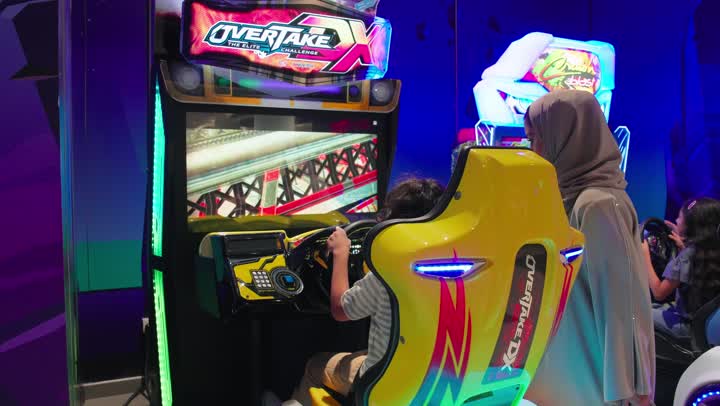 أجواء ترفيهية داخل مركز ألعاب إلكترونية حديث، تصوير من الخلف لولد عربي خليجي اماراتي يجلس على كرسي ألعاب إلكترونية ويقوم بلعب لعبة سباق السيارات باستخدام جهاز محاكاة القيادة برفقة والدته ، قضاء وقت ممتع مع الأسرة، مفهوم الترفيه العائلي والتفاعل الأسري داخل صالة الألعاب