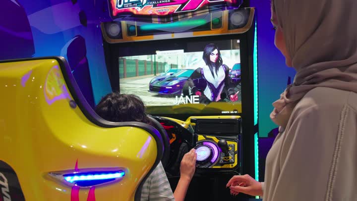 قضاء وقت ممتع مع الأسرة، أجواء ترفيهية داخل مركز ألعاب إلكترونية, تصوير من الخلف لطفل عربي خليجي اماراتي يرتدي الزي الكاجوال يقوم بلعب لعبة سباق سيارات إلكترونية برفقة والدته
