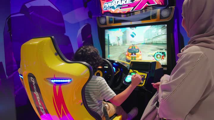 ام تقوم بالتصفيق لابنها بايماءات التشجيع والتحفيز، أجواء ترفيهية داخل مركز ألعاب إلكترونية, تصوير من الخلف لطفل عربي خليجي اماراتي يرتدي الزي الكاجوال يقوم بلعب لعبة سباق سيارات إلكترونية برفقة والدته, قضاء وقت ممتع مع الأسرة