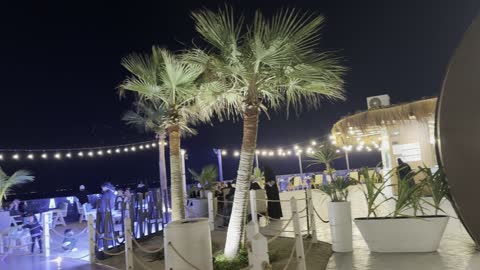 لقطة ليلية لمنطقة جلوس بأضواء متدلية وأشجار صغيرة، معالم سياحية مشهورة، الواجهة البحرية السياحية في المملكة العربية السعودية بمدينة الجبيل الصناعية ، كورنيش الجبيل، مجموعة من المطاعم والمقاهي المطلة على البحر