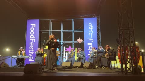 An Egyptian company specialized in dates and other food products, a traditional musical band on a lit stage, the Ithra Food Festival from December 22 to January 1, 2026, at the King Abdulaziz Center for World Culture (Ithra) in Dhahran, Saudi Arabia, a unique design resembling pebbles.