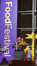 موسيقي يعزف على غيتار بزي عربي، فرقة موسيقية تقليدية على خشبة مسرح مضاءة، مهرجان إثراء للطعام من ٢٢ ديسمبر حتى ع يناير ٢٠٢٦، مركز الملك عبد العزيز للثقافة العالمية (إثراء) في الظهران في المملكة العربية السعودية، تصميم فريد يشبه الحصى