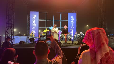 وجود الجماهيرالمحبة للفن والموسيقى العربية، فرقة موسيقية تقليدية على خشبة مسرح مضاءة، مهرجان إثراء للطعام من ٢٢ ديسمبر حتى ع يناير ٢٠٢٦، مركز الملك عبد العزيز للثقافة العالمية (إثراء) في الظهران في المملكة العربية السعودية، تصميم فريد يشبه الحصى