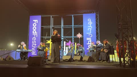 فرقة موسيقية تقليدية على خشبة مسرح مضاءة، عرض موسيقي حي في مهرجان للطعام، مهرجان إثراء للطعام من ٢٢ ديسمبر حتى ع يناير ٢٠٢٦، مركز الملك عبد العزيز للثقافة العالمية (إثراء) في الظهران في المملكة العربية السعودية، تصميم فريد يشبه الحصى