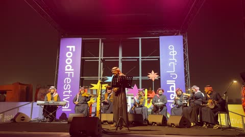 مغني يؤدي عرض حي على خشبة المسرح محاط بموسيقيين، فرقة موسيقية تقليدية على خشبة مسرح مضاءة، مهرجان إثراء للطعام من ٢٢ ديسمبر حتى ع يناير ٢٠٢٦، مركز الملك عبد العزيز للثقافة العالمية (إثراء) في الظهران في المملكة العربية السعودية، تصميم فريد يشبه الحصى