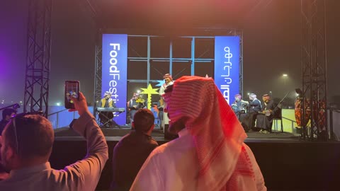 وجود الجماهيرالمحبة للفن والموسيقى العربية، فرقة موسيقية تقليدية على خشبة مسرح مضاءة، مهرجان إثراء للطعام من ٢٢ ديسمبر حتى ع يناير ٢٠٢٦، مركز الملك عبد العزيز للثقافة العالمية (إثراء) في الظهران في المملكة العربية السعودية، تصميم فريد يشبه الحصى