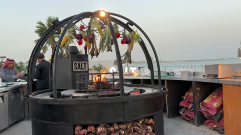 The circular wood-burning fireplace at the Salt of the Sea on the Corniche of Khobar in the Eastern Province, Saudi Arabia, offers recreational activities and events, a family-friendly coastal beach destination, tourist restaurants and cafes, a calm atmosphere, and a pleasant relaxation experience, with seating overlooking the sea.