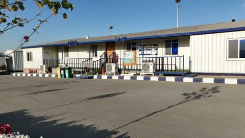 مبنى جاهز خارجي أبيض وسقف أزرق، مصنع متخصص في أعمال الخرسانة الجاهزة والأساسات الميكانيكية ، خلط مكونات الخرسانة ، عجينة اسمنتية للبناء والانشاء ، اماكن انشائية، أكوام كبيرة من المواد الخام الحصى، الرمل أو الركام مخزنة تحت هياكل