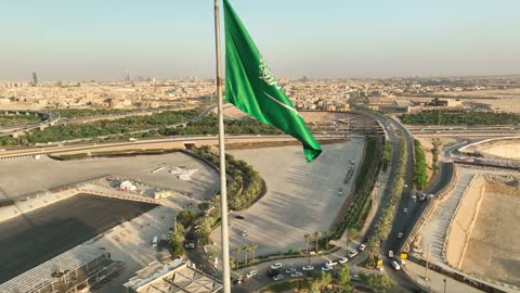 The flag of the Kingdom is waving over Diriyah, the development project of historic Diriyah in the city of Riyadh, Saudi Arabia, the heritage village and archaeological buildings, a center and a famous cultural and historical tourist landmark in the evening, the illuminated Al-Bujairi Park and overlook during the day.