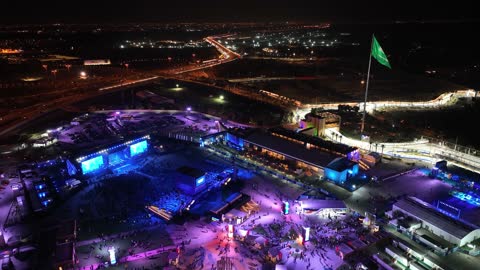 A green flag fluttering on a pole, the famous roads, streets, and landmarks of Jeddah, drone footage of a road in the city of Jeddah in the Kingdom of Saudi Arabia at night, car traffic, towers and residential buildings, a large number of people in the area.