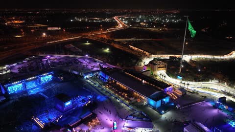 A green flag fluttering on a pole, the famous roads, streets, and landmarks of Jeddah, drone footage of a road in the city of Jeddah in the Kingdom of Saudi Arabia at night, car traffic, towers and residential buildings, a large number of people in the area.