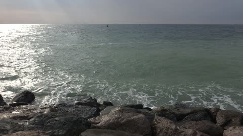 غروب الشمس فوق البحر في نخلة جميرا. أمواج تتلاطم على الصخور على شاطئ دبي الصخري. أمواج المحيط تتلاطم على الصخور المغطاة بالطحالب عند غروب الشمس مع سماء زرقاء صافية.