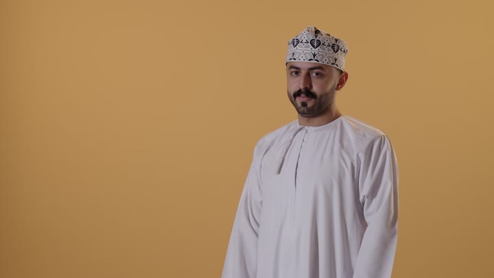 الوقوف باستقامة وثقه بالنفس، مفهوم الاناقه و الرجولة، بورتريه لشاب عربي خليجي عماني يرتدي الدشداشة و الكمة يقف بأيدي متقاطعه ينظر إلى الكاميرا بتعابير الجدية، الاهتمام بالمظهر الخارجي، خلفية برتقالية