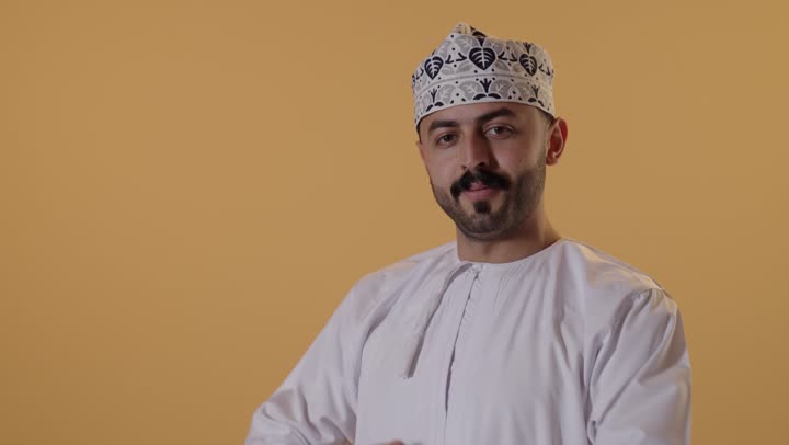 مفهوم الاناقه و الرجولة، الوقوف باستقامة وثقه بالنفس، بورتريه مقرب لشاب عربي خليجي عماني يرتدي الدشداشة و الكمة يقف بأيدي متقاطعه ينظر إلى الكاميرا بتعابير الجدية، الاهتمام بالمظهر الخارجي، خلفية برتقالية