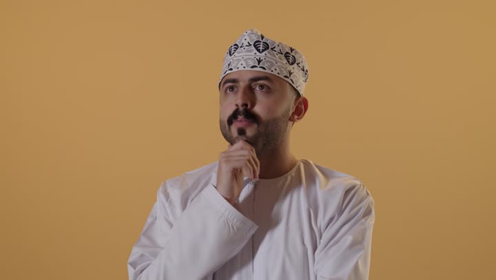 التركيز وشرود الذهن والتمعن، التأمل والتخطيط للمستقبل ، إيجاد حل لمشكلة ما، بورتريه مقرب لرجل عربي عماني يرتدي الدشداشة والكمه يضع يده على ذقنه و ينظر إلى شيء ما بإيماءات التفكير،خلفية برتقالية