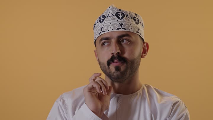 التأمل والتخطيط للمستقبل، التركيز وشرود الذهن والتمعن ، إيجاد حل لمشكلة ما، بورتريه مقرب لرجل عربي عماني يرتدي الدشداشة والكمه يضع يده على ذقنه و ينظر إلى شيء ما بإيماءات التفكير،خلفية برتقالية