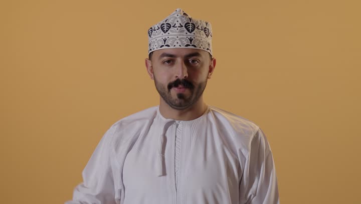 إنتظار مكالمة وإستخدام الميكروفون، بورتريه مقرب لشاب عربي خليجي عماني يرتدي الدشداشة والكمه يرفع يده بإيماءات الإتصال وإجراء مكالمة هاتفية، تعابير الفرح والسرور،  خلفية برتقالية