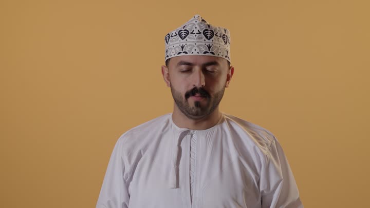 آلام الشقيقة والصداع النصفي، الشعور بالمرض وألم الرأس، الحاجه إلى العلاج والمساعده, بورتريه  مقرب لرجل عربي خليجي عماني يرتدي الدشداشة و الكمة يضع يديه على رأسه بإيماءات الانزعاج والتعب، خلفية برتقالية