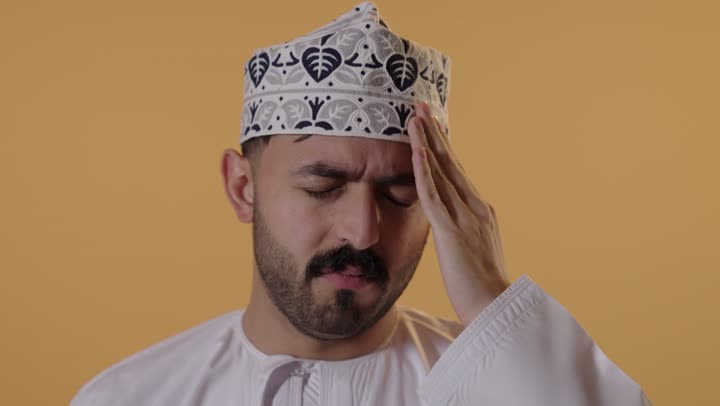 الشعور بالمرض وألم الرأس, آلام الشقيقة والصداع النصفي، الحاجه إلى العلاج والمساعده, بورتريه  مقرب لرجل عربي خليجي عماني يرتدي الدشداشة و الكمة يضع يديه على رأسه بإيماءات الانزعاج والتعب، خلفية برتقالية