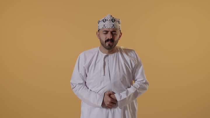 عسر الهضم والشعور بحرقة في المعدة، بورتريه لشاب عربي خليجي عماني يرتدي الدشداشة و الكمة ينحني ويضع يده على بطنه بإيماءات الألم، الإحساس بالمغص و التعب والإرهاق, طلب المساعده للذهاب الى الطبيب، الحاجة للدواء ووضع خطة علاجية، خلفية برتقالية