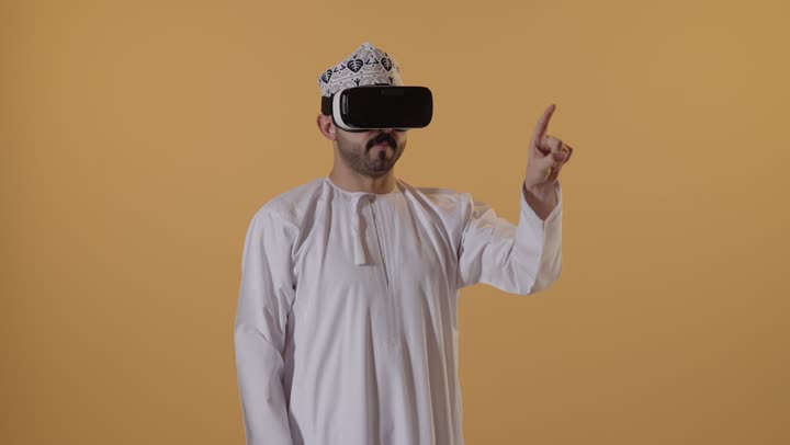 رؤية المشاهد الخيالية، التفاعل والاندماج مع الواقع الافتراضي، الاشارة بإصبع السبابة إلى شيء ما، استخدام الأجهزة التقنية الحديثة، بورتريه لرجل عربي خليجي عماني يرتدي الدشداشة والكمة يضع نظارات الفي آر، قضاء أوقات الفراغ بشكل ممتع، خلفية برتقالية