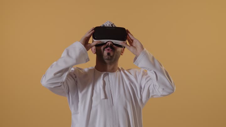 التطور التقني والتكنولوجي, المتعة و التسلية بالمشاهدة عبر نظارة الواقع الافتراضي، بورتريه مقرب لرجل عربي خليجي عماني يرتدي الدشداشه والكمه ينظر من خلال نظارات ثلاثية الأبعاد (الفي ار), الاندماج ومحاكاة العالم الخيالي والنظر الى الافلام الواقعيه، خلفيه برتقاليه