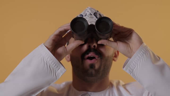 رؤية الاشياء البعيدة و تقريبها، حب الاستكشاف و المغامرة, بورتريه مقرب لرجل عربي خليجي عماني يرتدي الدشداشة و الكمة يحمل بيده المنظار ثنائي العينين ينظر الى شيء ما بايماءات السرور والذهول، مفهوم النظرة المستقبلية، خلفية برتقالية