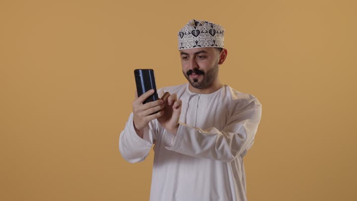 استخدام الأجهزة التقنية الحديثة للتسلية و الترفيه، تصفح وسائل التواصل الاجتماعي عبر الجوال, بورتريه لرجل عربي خليجي عماني يرتدي الدشداشة و الكمة يمسك الهاتف المحمول بيده, خلفية برتقالية