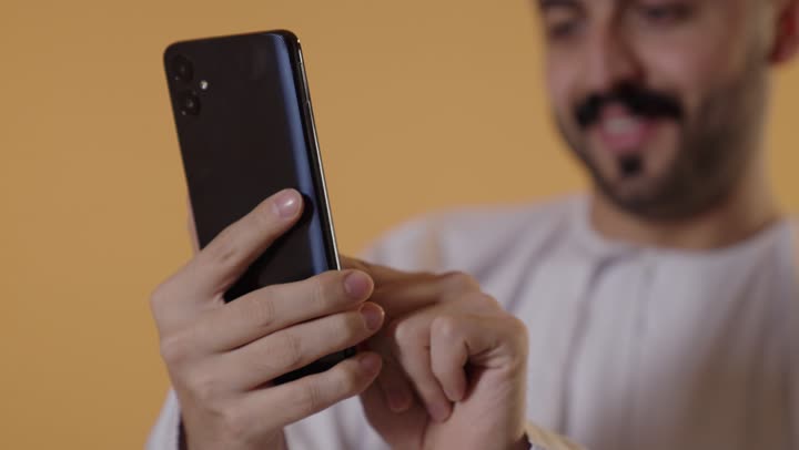 تصفح وسائل التواصل الاجتماعي عبر الجوال، استخدام الأجهزة التقنية الحديثة للتسلية و الترفيه, بورتريه مقرب لرجل عربي خليجي عماني يرتدي الدشداشة و الكمة يمسك الهاتف المحمول بيده, خلفية برتقالية